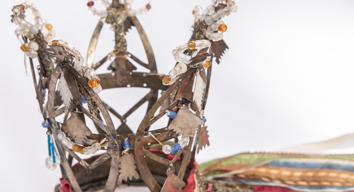 Brudekrone i metall med fem spir. Krona er pyntet med metallheng og glassperler. Det er påsydd valk nederst, med brodert bånd og metallknipling rundt valken.

Bak på det broderte båndet rundt valken, er det festet bånd. På midten er det et bredt bånd i rødt med metallkniplinger, med brede bånd med lys bunn og med en rød kant vendt ut fra den røde midtbåndet. Metallkniplinger er festet til de tre brede båndene. Deretter fire smalere bånd på hver side av de brede båndene. Disse er festet samlet med en liten sløyfe på det bredeste der det er festet på valken. 

En plastkam er påsydd valken sammen med to hvite, nyere bånd. Antagelig gjort i nyere tid for å feste krona og knyte under haka.