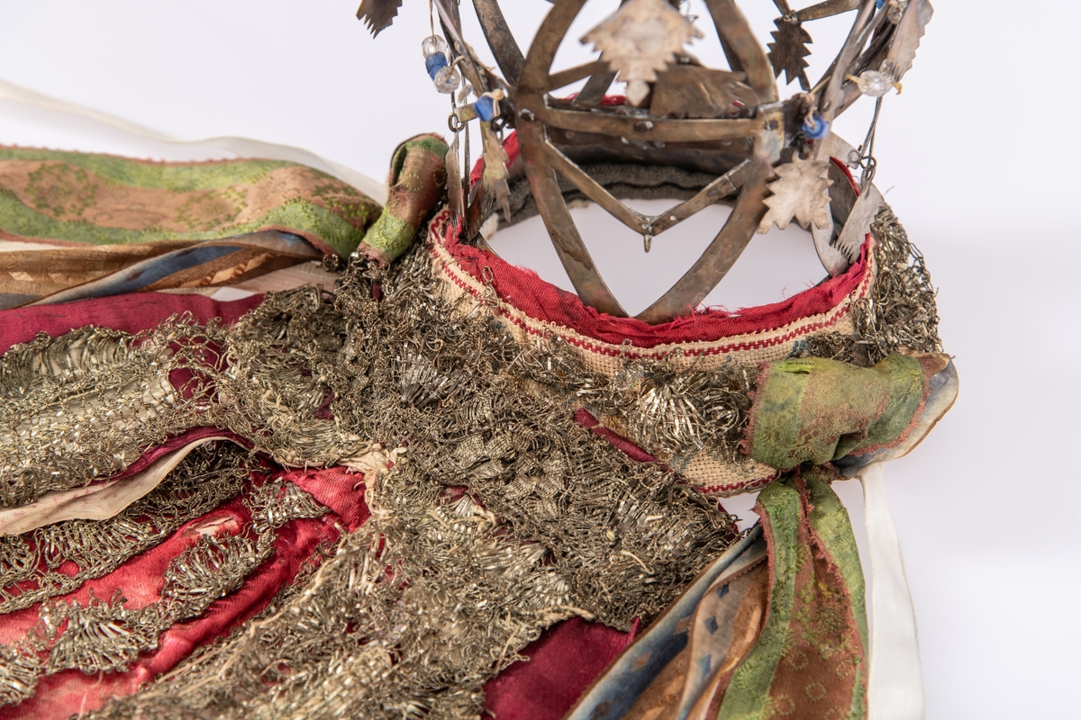 Brudekrone i metall med fem spir. Krona er pyntet med metallheng og glassperler. Det er påsydd valk nederst, med brodert bånd og metallknipling rundt valken.

Bak på det broderte båndet rundt valken, er det festet bånd. På midten er det et bredt bånd i rødt med metallkniplinger, med brede bånd med lys bunn og med en rød kant vendt ut fra den røde midtbåndet. Metallkniplinger er festet til de tre brede båndene. Deretter fire smalere bånd på hver side av de brede båndene. Disse er festet samlet med en liten sløyfe på det bredeste der det er festet på valken. 

En plastkam er påsydd valken sammen med to hvite, nyere bånd. Antagelig gjort i nyere tid for å feste krona og knyte under haka.