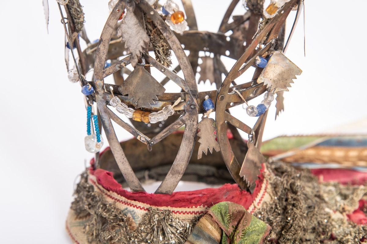 Brudekrone i metall med fem spir. Krona er pyntet med metallheng og glassperler. Det er påsydd valk nederst, med brodert bånd og metallknipling rundt valken.

Bak på det broderte båndet rundt valken, er det festet bånd. På midten er det et bredt bånd i rødt med metallkniplinger, med brede bånd med lys bunn og med en rød kant vendt ut fra den røde midtbåndet. Metallkniplinger er festet til de tre brede båndene. Deretter fire smalere bånd på hver side av de brede båndene. Disse er festet samlet med en liten sløyfe på det bredeste der det er festet på valken. 

En plastkam er påsydd valken sammen med to hvite, nyere bånd. Antagelig gjort i nyere tid for å feste krona og knyte under haka.