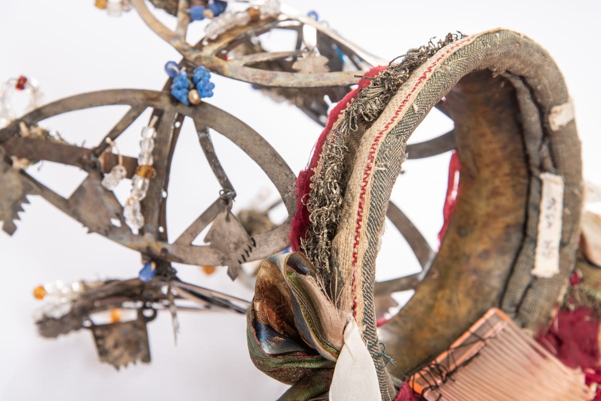 Brudekrone i metall med fem spir. Krona er pyntet med metallheng og glassperler. Det er påsydd valk nederst, med brodert bånd og metallknipling rundt valken.

Bak på det broderte båndet rundt valken, er det festet bånd. På midten er det et bredt bånd i rødt med metallkniplinger, med brede bånd med lys bunn og med en rød kant vendt ut fra den røde midtbåndet. Metallkniplinger er festet til de tre brede båndene. Deretter fire smalere bånd på hver side av de brede båndene. Disse er festet samlet med en liten sløyfe på det bredeste der det er festet på valken. 

En plastkam er påsydd valken sammen med to hvite, nyere bånd. Antagelig gjort i nyere tid for å feste krona og knyte under haka.