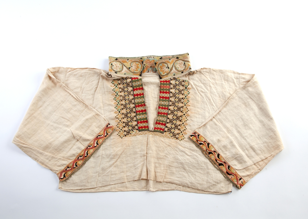 Skjorte fra Sauland, Hjartdal. Broderi på bolen uttrekksøm, klostersøm og korssting. Halslinning og slengerm sydd i plattsøm 
Halskvarde (halslinning) mål:  7,7cm.x57cm ca. Broderi 6,8cm.
Broderi på skulder ca 5,5x6,5cm. 
Brodert bord på slengerm 4cm., avslutning knapphullskant og attersting.