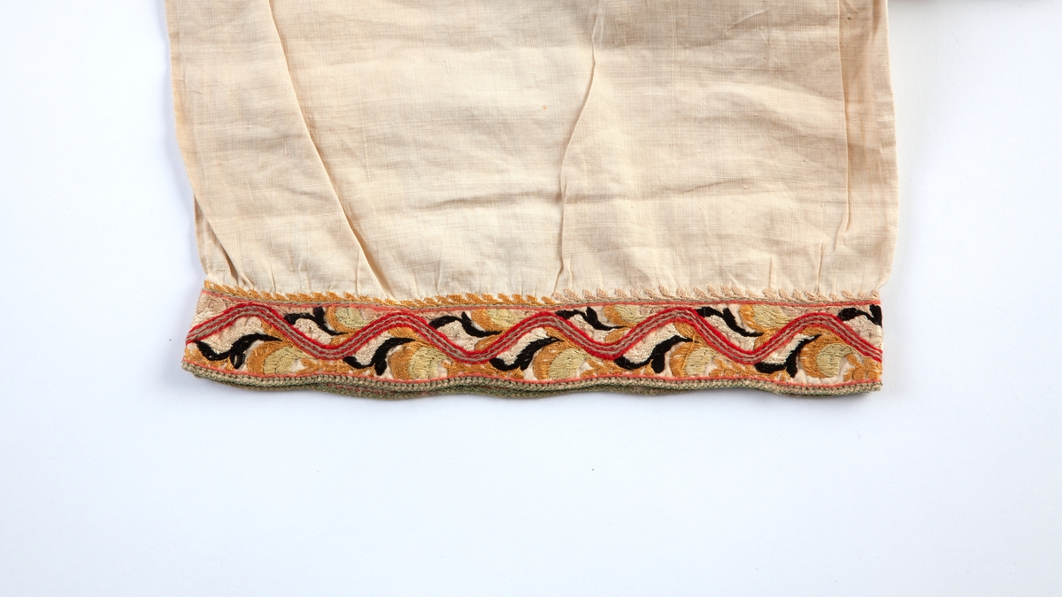 Skjorte fra Sauland, Hjartdal. Broderi på bolen uttrekksøm, klostersøm og korssting. Halslinning og slengerm sydd i plattsøm 
Halskvarde (halslinning) mål:  7,7cm.x57cm ca. Broderi 6,8cm.
Broderi på skulder ca 5,5x6,5cm. 
Brodert bord på slengerm 4cm., avslutning knapphullskant og attersting.