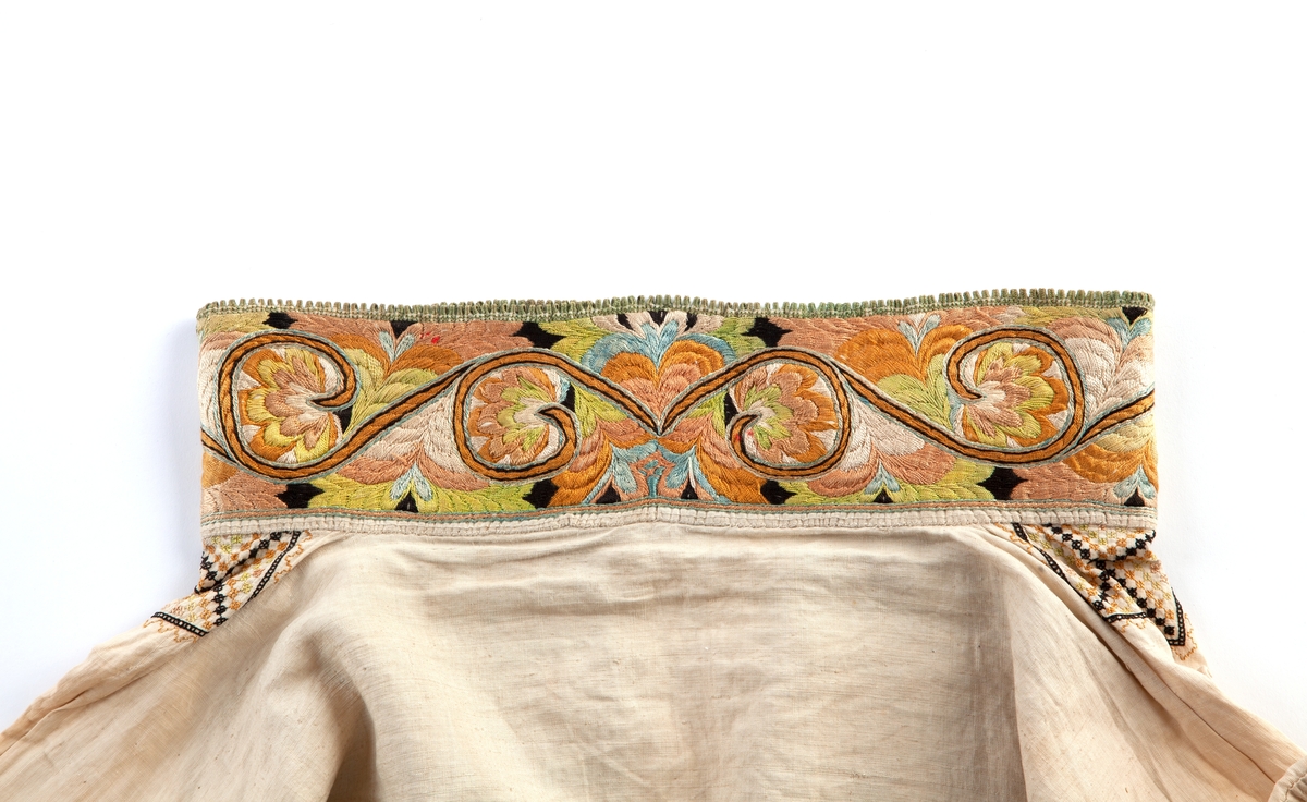 Skjorte fra Sauland, Hjartdal. Broderi på bolen uttrekksøm, klostersøm og korssting. Halslinning og slengerm sydd i plattsøm 
Halskvarde (halslinning) mål:  7,7cm.x57cm ca. Broderi 6,8cm.
Broderi på skulder ca 5,5x6,5cm. 
Brodert bord på slengerm 4cm., avslutning knapphullskant og attersting.