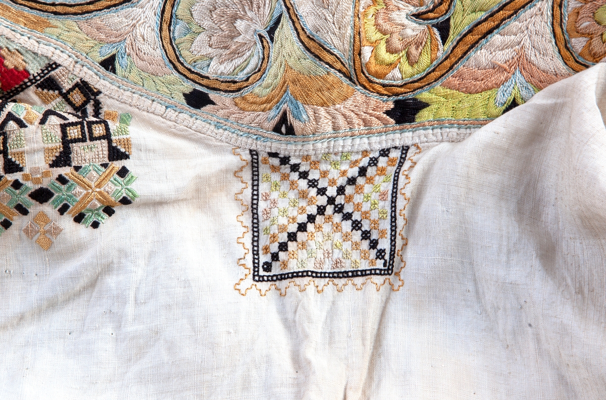 Skjorte fra Sauland, Hjartdal. Broderi på bolen uttrekksøm, klostersøm og korssting. Halslinning og slengerm sydd i plattsøm 
Halskvarde (halslinning) mål:  7,7cm.x57cm ca. Broderi 6,8cm.
Broderi på skulder ca 5,5x6,5cm. 
Brodert bord på slengerm 4cm., avslutning knapphullskant og attersting.