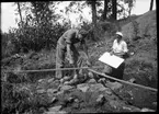 En stenpackning dokumenteras i samband med arkeologiska undersökningar vid Rumlaborg i Huskvarna sommaren 1934. Sittande med ritbrädet är fil.stud. Anders Lindahl. Han får hjälp med mätningen av skoleleven Gunnar Hellman. Anders Lindahl var vid undersökningarna mellan 1933-1938 projektledaren Georg Sahlströms närmaste man. Gunnar Hellman hade samma roll vid undersökningarna 1939 och 1942.