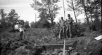 Tre män fotograferade i samband med den arkeologiska undersökningen vid Rumlaborg i Huskvarna sommaren 1934. I främre delen av bilden syns en framgrävd stenpackning. Personen till vänster är fil. stud. Anders Lindahl som står vid ett avvägningsinstrument. Personen som håller i lattan är troligen skoleleven Gunnar Hellman medan personen med spaden inte har kunnat identifieras.