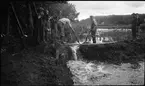 Några män och skolpojkar står vid en fördämning mellan Huskvarnaån och den inre vallgraven vid Rumlaborg i Huskvarna. Fotot togs i samband med arkeologiska undersökningar vid borgen sommaren 1935.