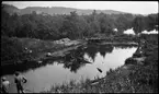 Två män står vid den inre vallgraven vid Rumlaborg i Huskvarna i samband med arkeologiska undersökningar sommaren 1935. Vattennivån i vallgraven är sänkt med hjälp av en pump och mot Huskvarnaån har en fördämning byggts. Centralt i bilden syns medeltida stockrester.