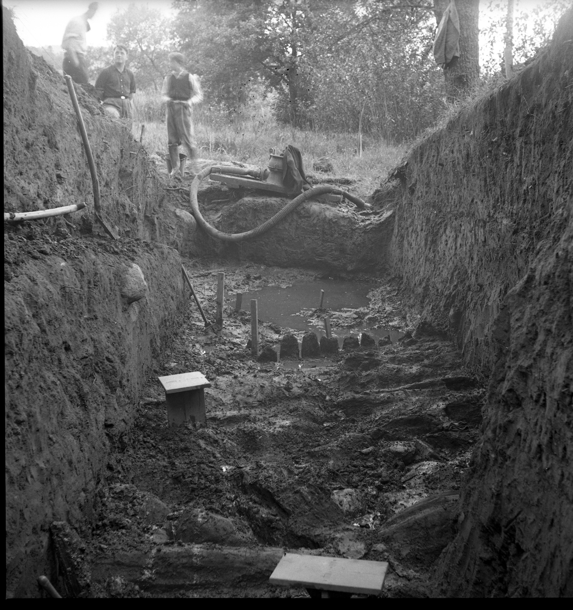 Schakt D genom den södra delen av borgkullen vid Rumlaborg i Huskvarna i samband med arkeologiska undersökningar sommaren 1938. Fotot är taget mot söder och i förgrunden kan stockrester ses, lite längre fram även rester av en pålrad. I bakgrunden står tre män, vid sidan av pumpen som användes för att hålla schaktet någorlunda torrt. Rester efter pålrader påträffades även vid andra delar längs med borgkullens kant i samband med de arkeologiska undersökningarna, som bedrevs av Huskvarna hembygdsförening mellan åren 1931-1942.