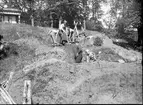 Fyra skolpojkar och en man från Huskvarna stad gräver i schakt G vid sydvästra sluttningen av borgkullen vid Rumlaborg i Huskvarna sommaren 1942. I bakgrunden syns lusthuset på kullens topp.