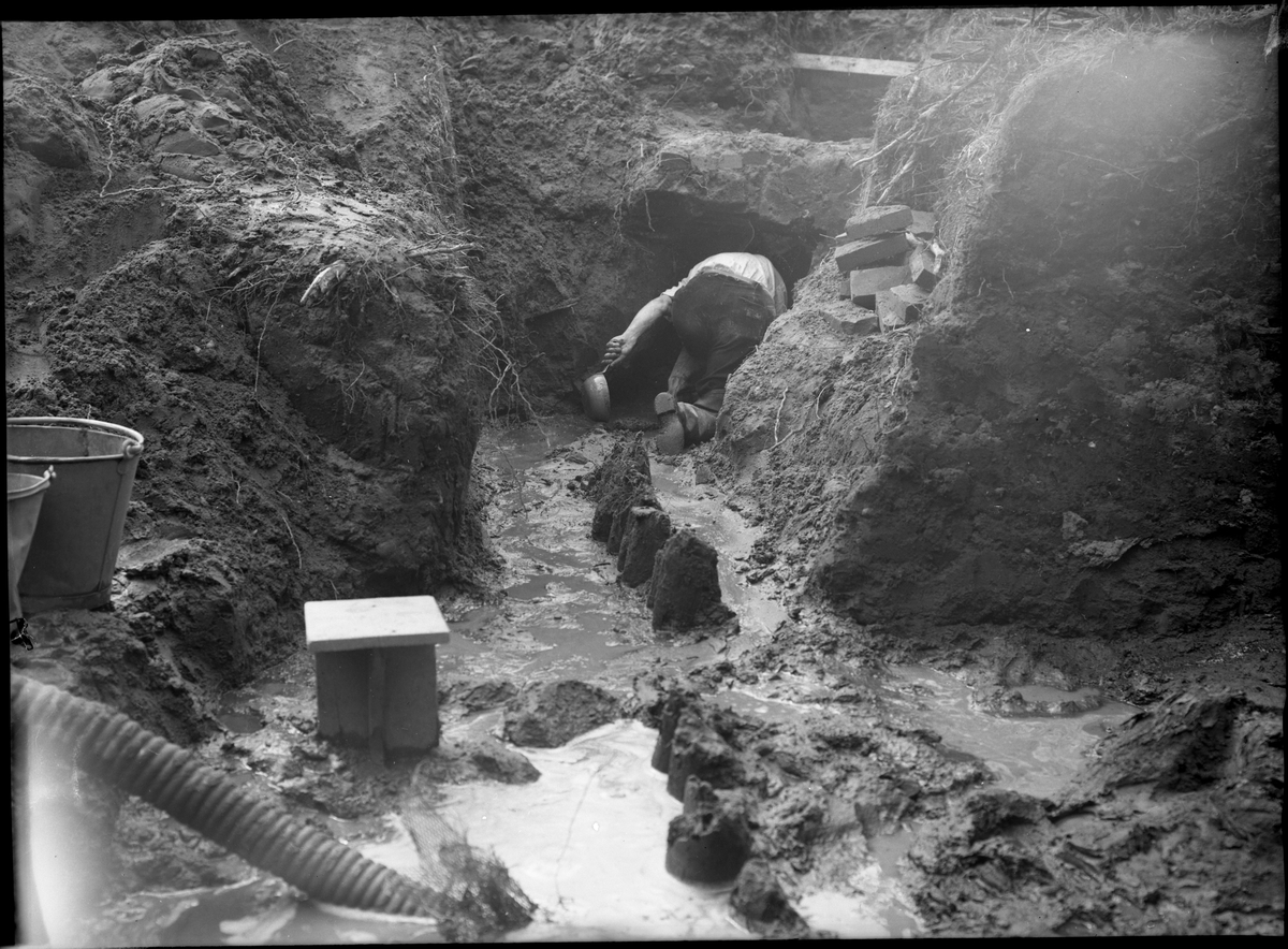 Schakt G utvidgas åt väster mot den gamla landsvägen. Foto taget i samband med de arkeologiska undersökningarna vid Rumlaborg i Huskvarna sommaren 1942. Centralt i bilden syns en rad med stolpar. För att få med fortsättningen av denna stolprad fattades beslut om att utvidga schaktet åt väster. I förgrunden syns röret till en diafragmadriven handpump som var nödvändig för att undersökningen av schaktet skulle gå att geomföra med tanke på inträngande vatten från Huskvarnaån.