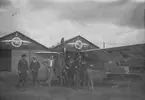 En flygare och personal vid Thulins flygskola står framför flygplan Thulin A, omkring 1915. Sju män. I bakgrunden hangarer.