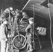 Flygplan Thulin A står i hangar på Thulins flygskola i Ljungbyhed, omkring 1915-1920. Två flygtekniker jobbar med flygplanet. På golvet står verkmästare Oskar Johansson till vänster och flygskolechefen Elis Nordqvvist.