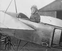 Snickaren Palle Mellblom iklädd flygdräkt sitter i flygplan Thulin A framför hangar på Thulins flygskola i Ljungbyhed, omkring 1915-1920.