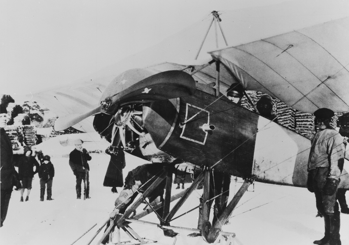 Flygplan Thulin D står på isen utanför Vasa, Finland, inför överlämnande till de finska styrkorna den 8/3 1918. I förarsitsen löjtnant Klintberg, i baksits en finsk kontrollant. Folksamling kring flygplanet.