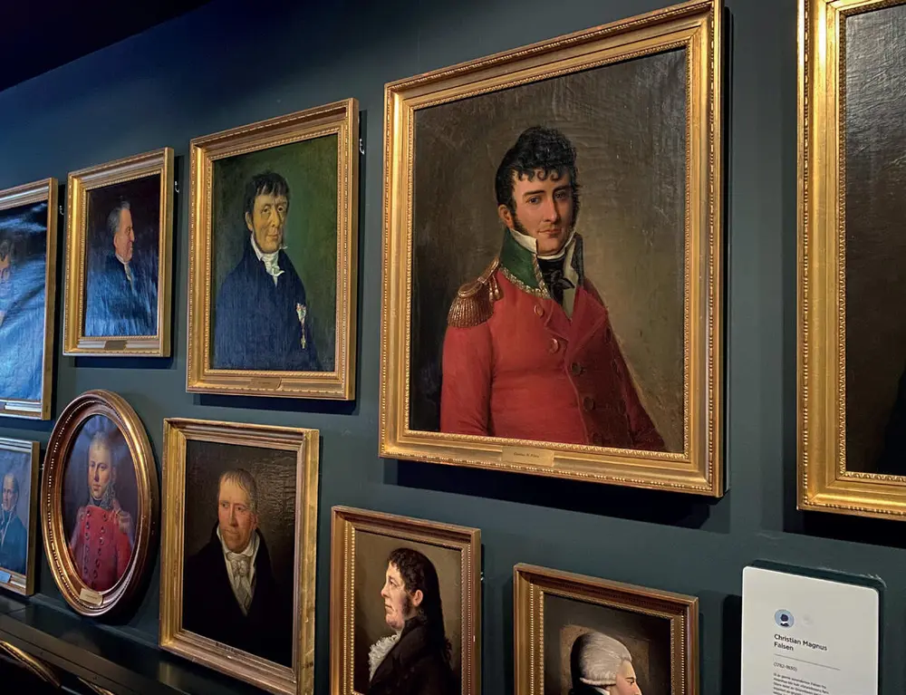 Et fotografi fra Eidsvollsgalleriet viser utsnitt av samlingen av portrettmalerier hengende på en vegg. I forgrunnen portrettet av Christian Magnus Falsen.