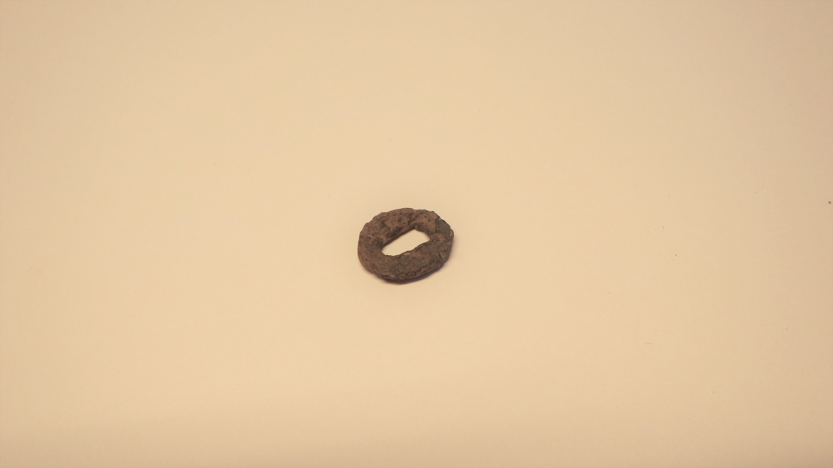 Oval plate med et rektangulært hull i.  Gjenstanden er trolig en del av skaftholken til en kniv. 