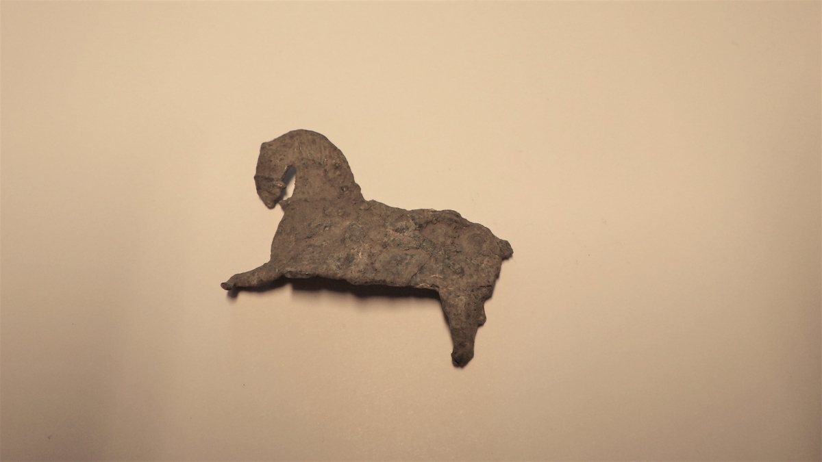 Metallplate fint formet som en hest med preg av seletøy. Den nedre delen av beina samt halen er i dag vekk, men deler av dekoren i form av seletøy, man og andre detaljer sees forsatt, selv om gjenstanden har en slitt overflate. Det samme preget sees på begge sider av platen. Gjenstanden er derfor trolig ikke en form for et beslag, men kanskje heller en leke ment for et barn. 