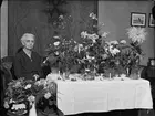 Kvinna i hemmiljö omgiven av blommor, Uppland 1948