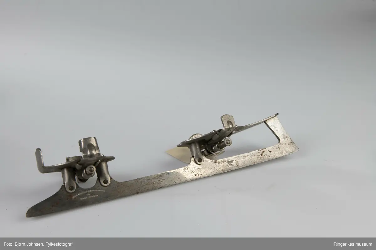 To skøyter i et par, ANT 1910-1920.Smalt og tynt skøytejern, avskåret spiss tupp  i bakre ende og skrådd avkuttet foran. Montert to flate plater til å sette støvlene på skøytejernet, det bakre er bøyd opp for plassering av helen. På undersiden av hver plate montert to tverstilte og bevegelige jern for feste av støvlen. De  reguleres med store muttere.