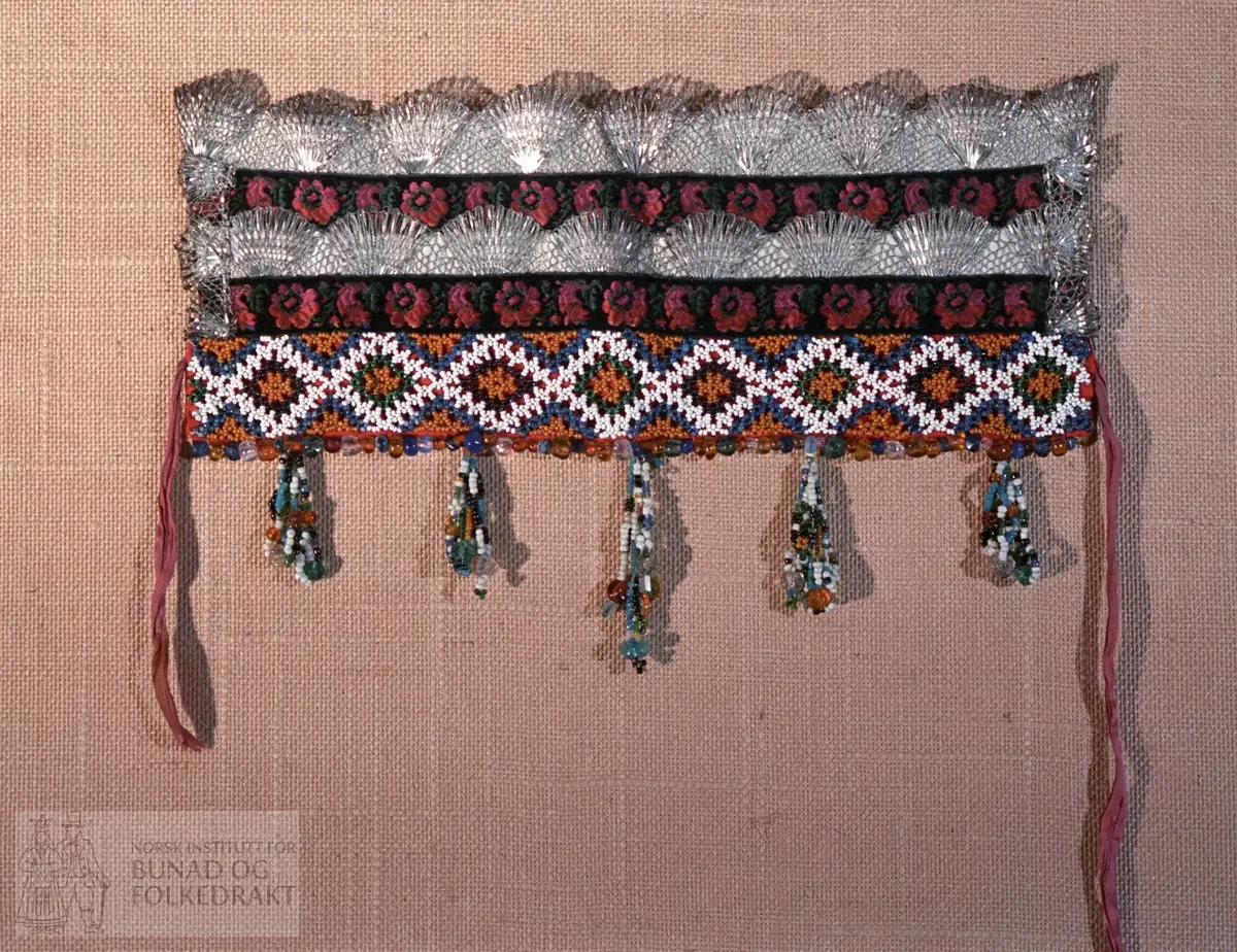 Bakpart rødt klede og kvitt bomullslerret med påsydd papp.   FORAN: Perlebroderi over kleden i kvite, gule, svarte, grønne og blåt perler.  De blå perlene er forholdsvis store.  Deretter (på lerrettsbunnen) påsydd nr 1: svart fløyelsband i lansert rosemønster.  Farger: grønt, rosa, rødt og gult.  Nr. 2:  bord med sølvknipling.   Nr. 3: svarte fløyelsband i lansert rosemønster.  Nr. 4: bord med sølvknipling.  I sidene - ca. midt på - påsydd et stykke med smalere sølvknipling.   Nederst - under perlebroderiet - perlerad med perleheng.  Her er det nytta perler i varierende størrelse i fargene kvitt, svart, grønt, gult og gjennomsiktige perler i fargene gult, grønt, blått og blankt.  Knytteband i bleikrosa silke - lengde 31 cm.  Se mål på kort.