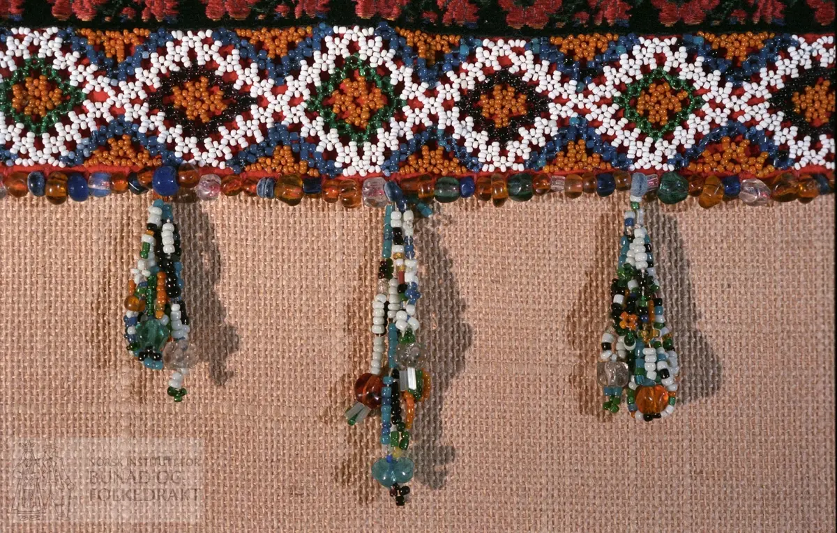 Bakpart rødt klede og kvitt bomullslerret med påsydd papp.   FORAN: Perlebroderi over kleden i kvite, gule, svarte, grønne og blåt perler.  De blå perlene er forholdsvis store.  Deretter (på lerrettsbunnen) påsydd nr 1: svart fløyelsband i lansert rosemønster.  Farger: grønt, rosa, rødt og gult.  Nr. 2:  bord med sølvknipling.   Nr. 3: svarte fløyelsband i lansert rosemønster.  Nr. 4: bord med sølvknipling.  I sidene - ca. midt på - påsydd et stykke med smalere sølvknipling.   Nederst - under perlebroderiet - perlerad med perleheng.  Her er det nytta perler i varierende størrelse i fargene kvitt, svart, grønt, gult og gjennomsiktige perler i fargene gult, grønt, blått og blankt.  Knytteband i bleikrosa silke - lengde 31 cm.  Se mål på kort.