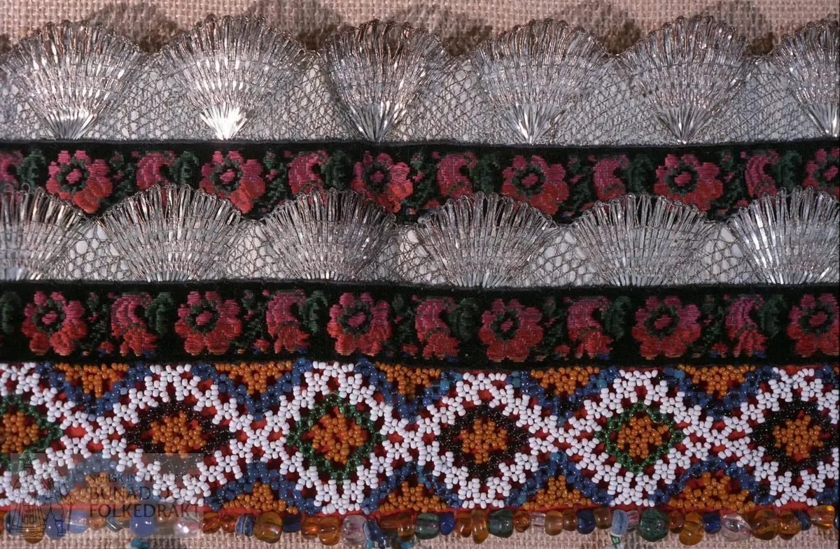 Bakpart rødt klede og kvitt bomullslerret med påsydd papp.   FORAN: Perlebroderi over kleden i kvite, gule, svarte, grønne og blåt perler.  De blå perlene er forholdsvis store.  Deretter (på lerrettsbunnen) påsydd nr 1: svart fløyelsband i lansert rosemønster.  Farger: grønt, rosa, rødt og gult.  Nr. 2:  bord med sølvknipling.   Nr. 3: svarte fløyelsband i lansert rosemønster.  Nr. 4: bord med sølvknipling.  I sidene - ca. midt på - påsydd et stykke med smalere sølvknipling.   Nederst - under perlebroderiet - perlerad med perleheng.  Her er det nytta perler i varierende størrelse i fargene kvitt, svart, grønt, gult og gjennomsiktige perler i fargene gult, grønt, blått og blankt.  Knytteband i bleikrosa silke - lengde 31 cm.  Se mål på kort.