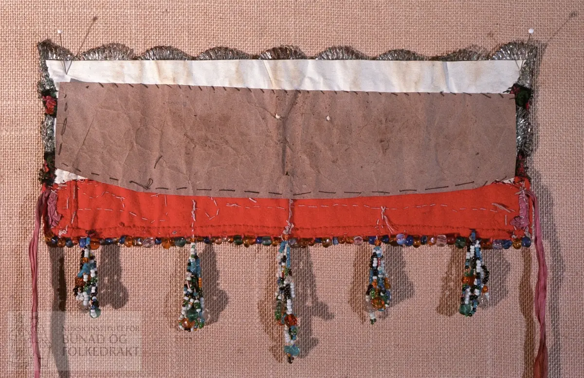 Bakpart rødt klede og kvitt bomullslerret med påsydd papp.   FORAN: Perlebroderi over kleden i kvite, gule, svarte, grønne og blåt perler.  De blå perlene er forholdsvis store.  Deretter (på lerrettsbunnen) påsydd nr 1: svart fløyelsband i lansert rosemønster.  Farger: grønt, rosa, rødt og gult.  Nr. 2:  bord med sølvknipling.   Nr. 3: svarte fløyelsband i lansert rosemønster.  Nr. 4: bord med sølvknipling.  I sidene - ca. midt på - påsydd et stykke med smalere sølvknipling.   Nederst - under perlebroderiet - perlerad med perleheng.  Her er det nytta perler i varierende størrelse i fargene kvitt, svart, grønt, gult og gjennomsiktige perler i fargene gult, grønt, blått og blankt.  Knytteband i bleikrosa silke - lengde 31 cm.  Se mål på kort.