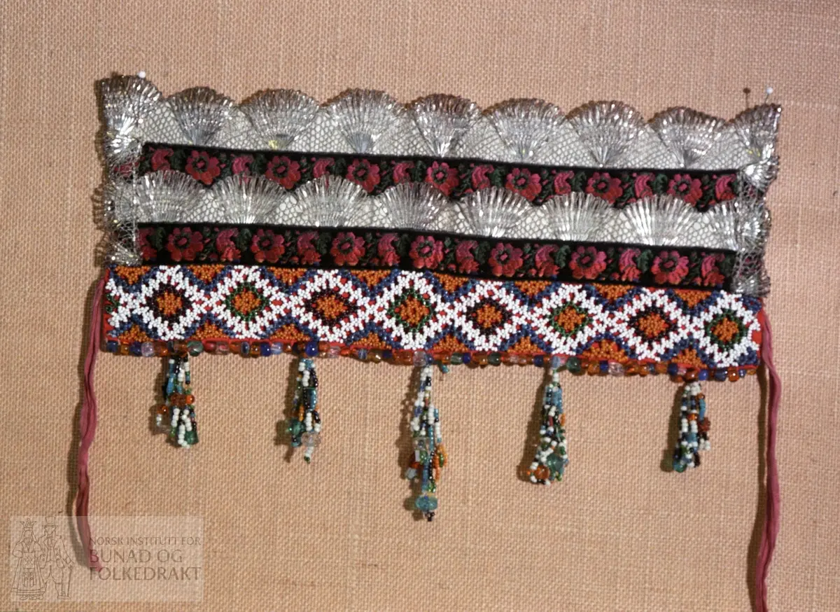 Bakpart rødt klede og kvitt bomullslerret med påsydd papp.   FORAN: Perlebroderi over kleden i kvite, gule, svarte, grønne og blåt perler.  De blå perlene er forholdsvis store.  Deretter (på lerrettsbunnen) påsydd nr 1: svart fløyelsband i lansert rosemønster.  Farger: grønt, rosa, rødt og gult.  Nr. 2:  bord med sølvknipling.   Nr. 3: svarte fløyelsband i lansert rosemønster.  Nr. 4: bord med sølvknipling.  I sidene - ca. midt på - påsydd et stykke med smalere sølvknipling.   Nederst - under perlebroderiet - perlerad med perleheng.  Her er det nytta perler i varierende størrelse i fargene kvitt, svart, grønt, gult og gjennomsiktige perler i fargene gult, grønt, blått og blankt.  Knytteband i bleikrosa silke - lengde 31 cm.  Se mål på kort.