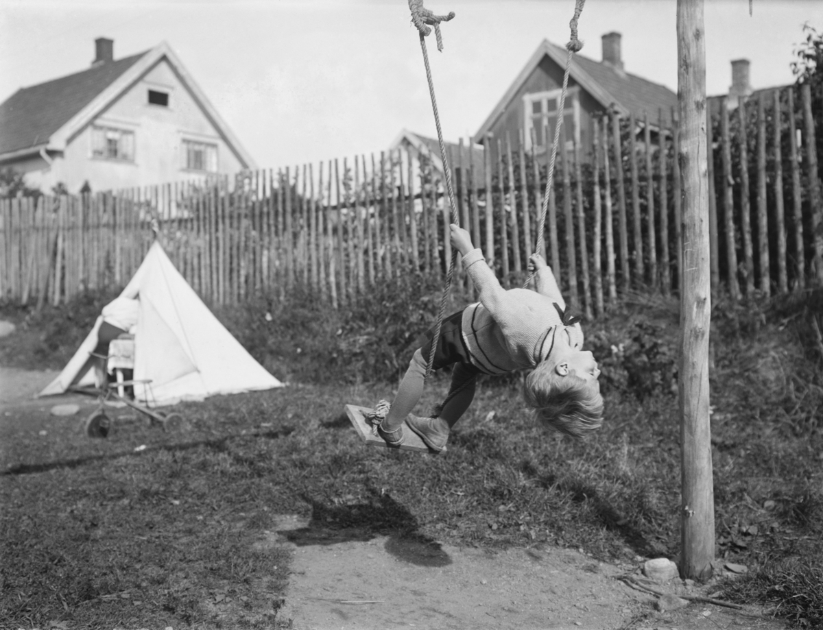 Liten gutt i huske. Gunnar Berge. Hus i bakgrunnen. Lite telt og tråsykkel.