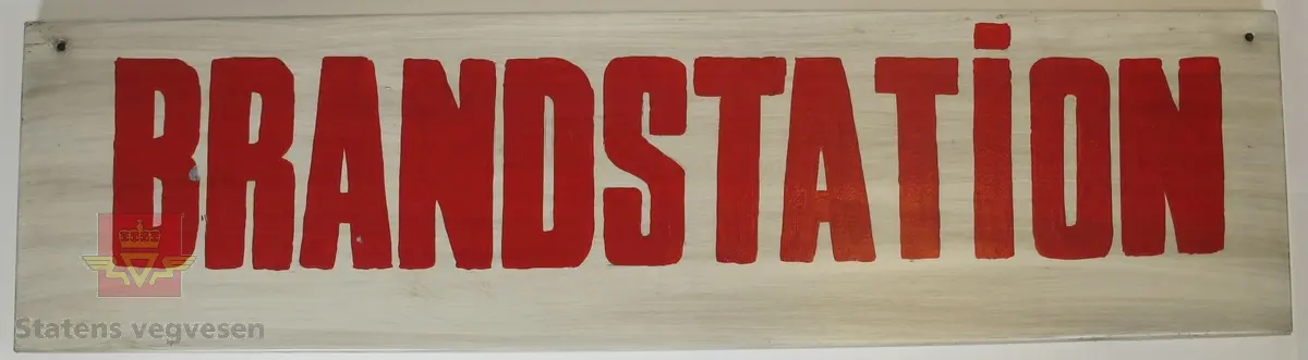 Rektangulært navneskilt lagd av metall, med teksten "BRANDSTATiON" påført med røde bokstaver på hvit bakgrunn. Det er laget et hull i hvert av de øverste hjørnene.