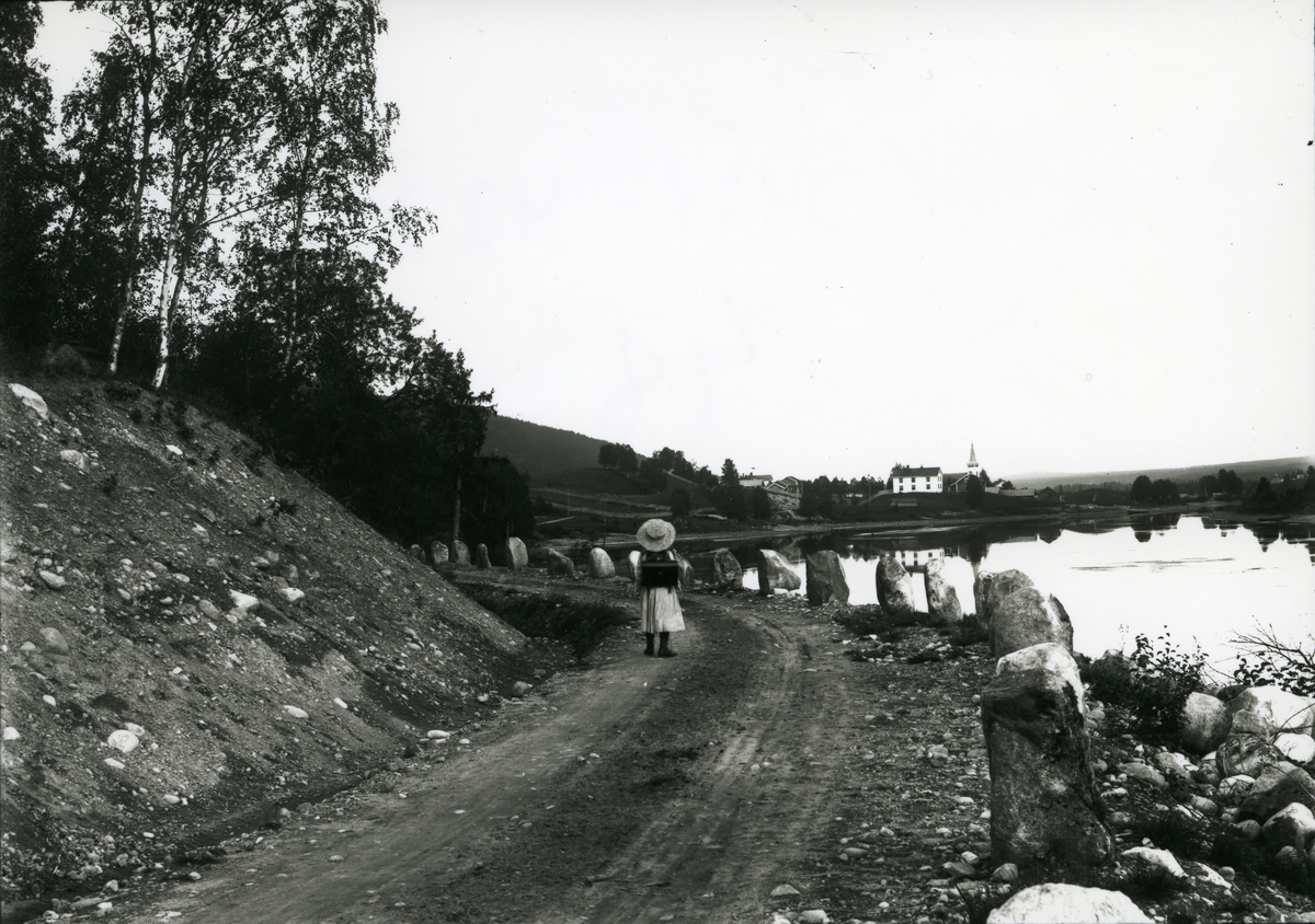 Østsiden fotografert nordfra. Nordby og kirken i bakgrunnen. Lita jente med hatt i forgrunnen. Elva til høyre.