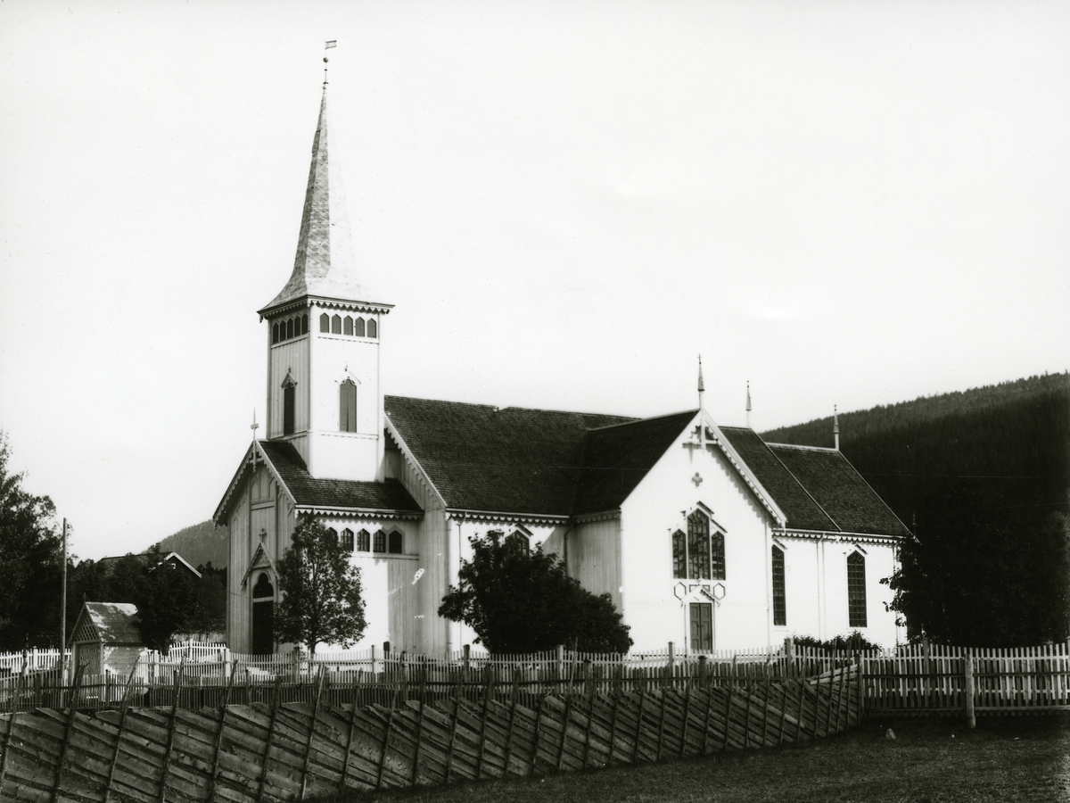 Trysil kirke fotografert fra sør-vest. 2 gjerder i forgrunnen. Port ved vestre inngangen.