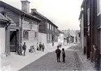 Vy längs Sankt Korsgatan i Linköping. Tiden är år 1900 och bebyggelsen speglar väl den enkelhet som länge var dominerande runt stadens omedelbara centrum. Närmast till vänster ses Gelbgjutaregården och till höger Bryggaregården, som oaktat sitt namn inrymt skiftande verksamheter över åren. Upplysningsvis är båda husen flyttade till friluftsmuseet Gamla Linköping.