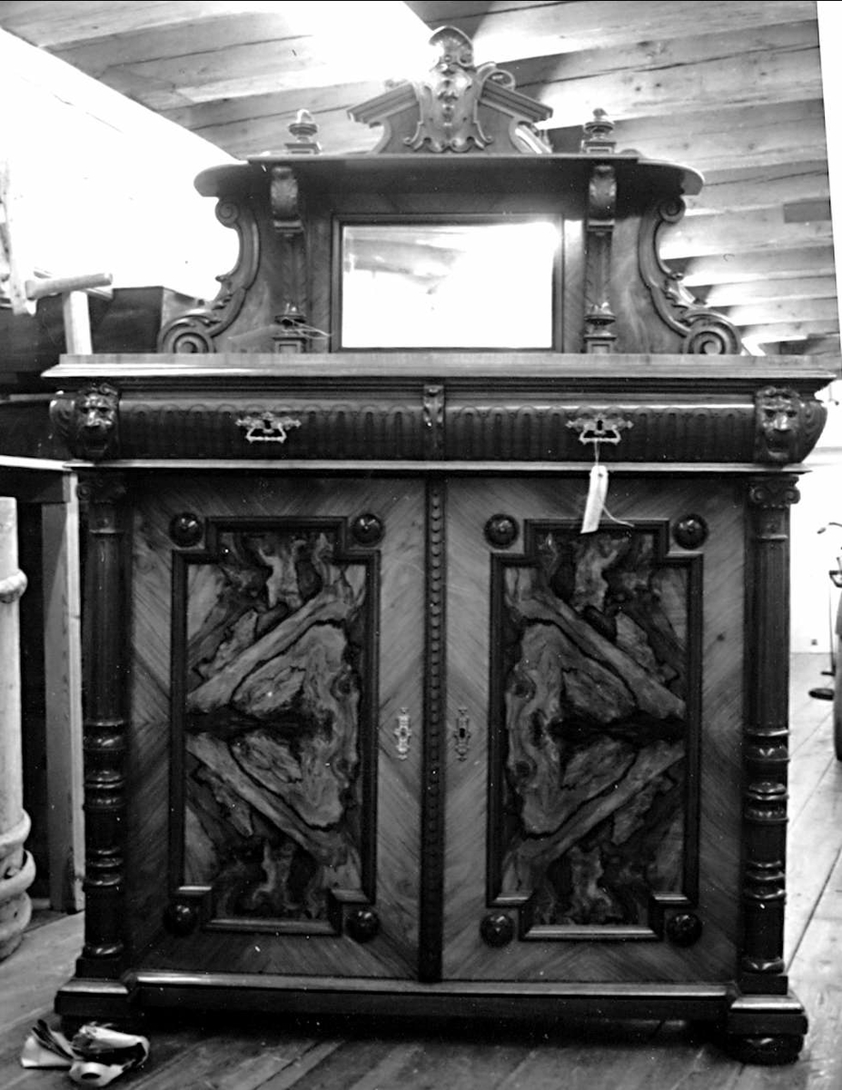 Skänk i nyrenässansstil. Av märkt polerat trä med rotfanér i speglarna och skivan. Två dörrar och över dem två lådor. På sidorna skulpturerade pilastrar med lejonhuvud upptill. Överstycke med rektangulär spegel och en bred hylla, dekor i form av voluter och bladverk och krönt med en snidad sköld. Mässingsbeslag. Nyckel märkt UM24492b. På baksidan skrivet med blyerts: Förfärdiga av Snickarmäst. J.A.Liljestrand År 1896.