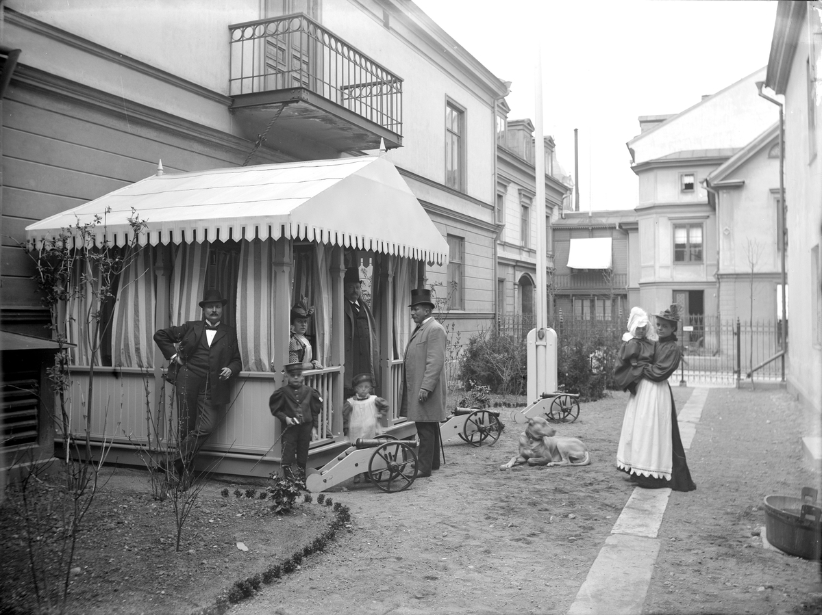 Gårdsinteriör från familjen Brobecks bostad i Stora hotellet i Linköping. Längst till vänster står familjefadern, grosshandlaren John Brobeck. På verandan sitter hans maka Hilda Ingeborg Lindqvist. Barnen är sannolikt parets tre föstfödda, John (f. 1892), Jim (f. 1893) och i barnflickans famn Jane (f. 1896). På bilden även postkontrollör Lind och trädgårdsdirektör Christian Mohr. Bild från något av 1800-talets sista år.