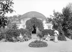 Från Wallenbergska trädgården i Linköping med ställets orginella paviljong. Byggnaden är beskriven av Wallenbergs systerdotter Ulla Linder (1873-1854), som i sin barndom ofta besökte platsen. Hon mindes att byggnaden innehöll inte mindre än fyra rum. Ett stort 8-kantigt rum i mitten, innanför detta ett mindre med ingång från mittsalen samt ett rum på vardera sida av mittenrummet men försedda med egna ingångar utifrån. Till det stora rummet kom man från trädgården via en närmare tre meter bred trappa och en liten loggia, vars tak bars upp av två doriska pelare. Ulla Linders beskrivning överenstämmer således väl med vad fotografiet visar, även om vildvinet tagit verandan i besittning. Vad vi emellertid inte ser är det stora rummets välvda innertak som Ulla beskriver var illusoriskt bemålat med valvbågar runt en stjärnhimmel där Aurora tronade bland molnen. Det inre rummet hade väggfasta soffor klädda med storblommig damast i rosenrött. Över sofforna hängde fyra stora litografier med porträtt av Sveriges alla konungar och drottningar samt i övrigt några namnkunniga personer ur den svenska historien. Fotografiet kan antas vara tillkommet 1898. Personerna har inte identifierats.