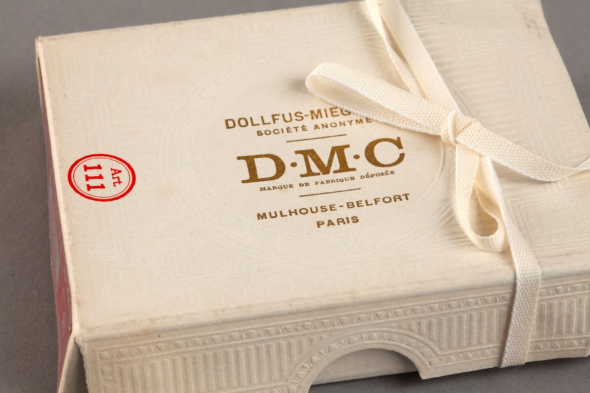 Förpackning i kartong. För garn. Vit. Jugendmönster i relief. Text i guld. På locket står: Dollfus-Mieg & Cie. Société anonyme DMC Marque de fabrique Déposés. Mulhouse Belfort Paris. Blå på insidan. Ett silkepapper ligger i. På sidan står: coton floche DMC qualité spéciale. 12 échev. Blanc neige. 8.