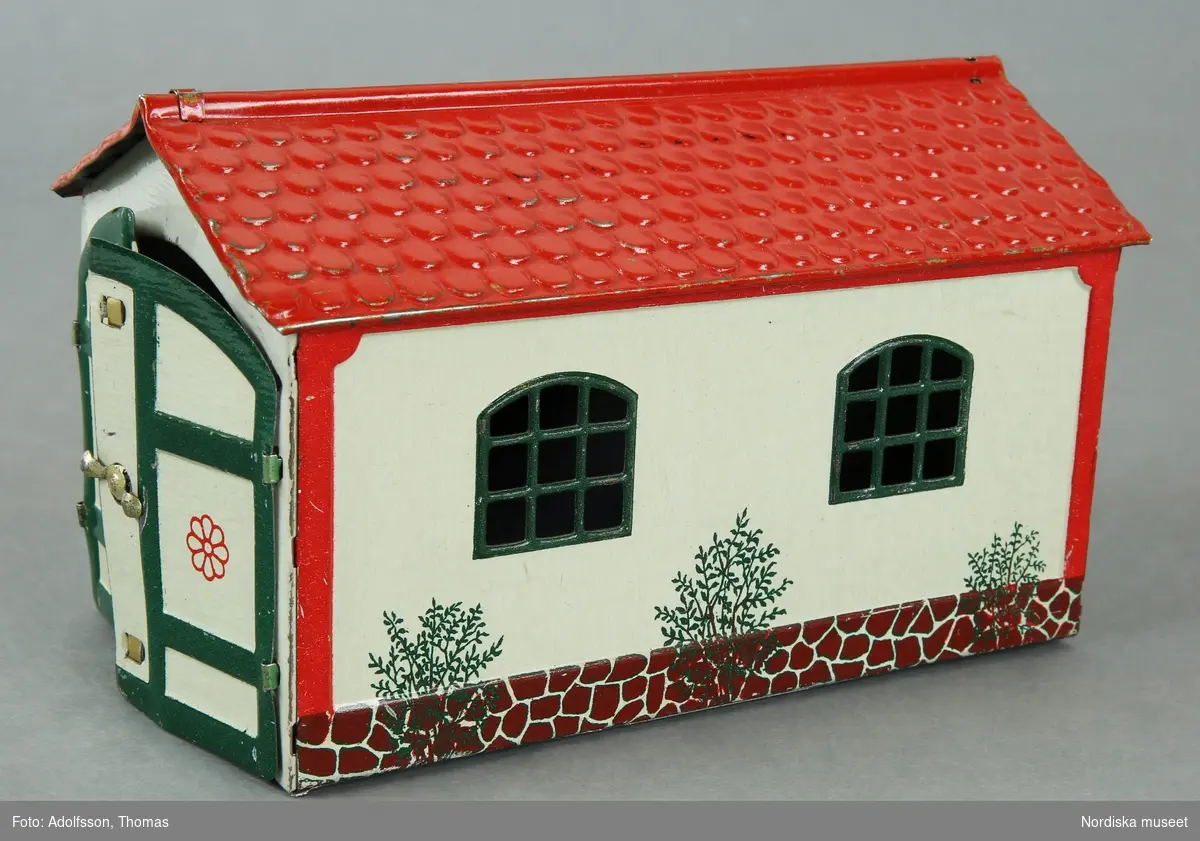 Inventering Sesam 1996-1999:
L 15,5 cm
B 4 cm
H 9,5 cm
Litograferat garage av plåt, gulvita väggar, rött tegeltak, gröna genombrutna fönster. Rörliga dörrar. Målad stenfot och buskar. Märkt: "LEHMANN / MADE IN GERMANY / 771 / D.R. G.M.". Botten genombruten.
K Berg 1972/= Kerstin Berg