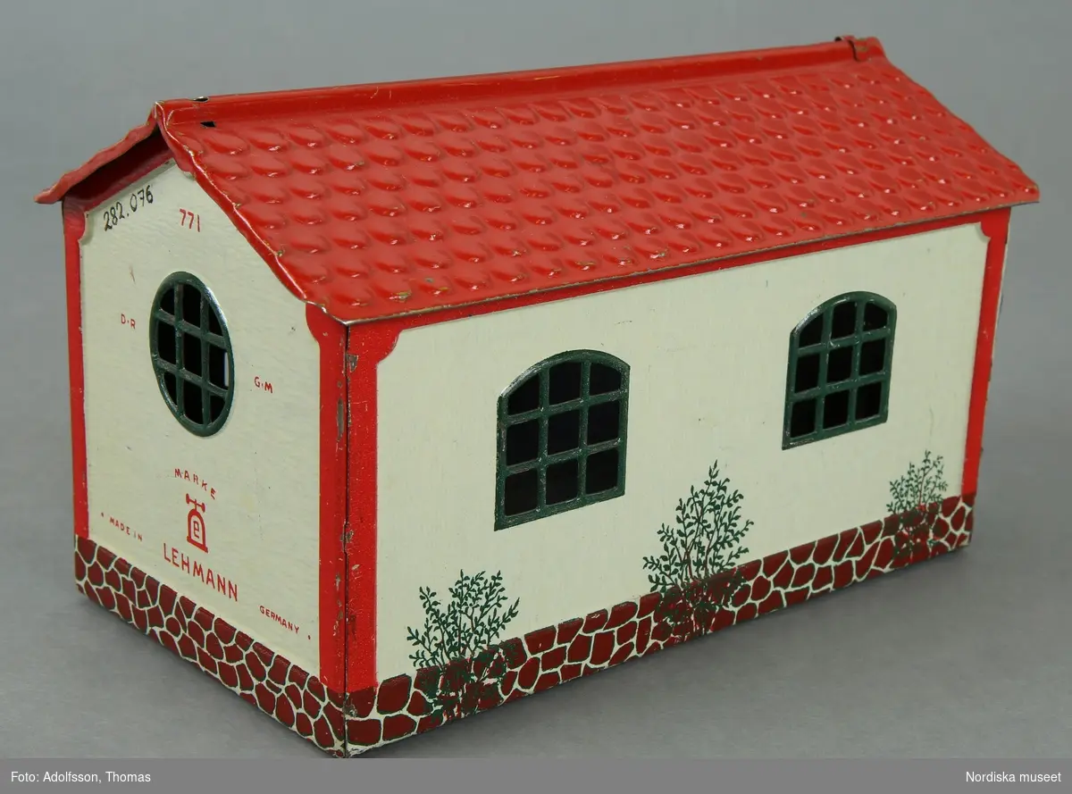 Inventering Sesam 1996-1999:
L 15,5 cm
B 4 cm
H 9,5 cm
Litograferat garage av plåt, gulvita väggar, rött tegeltak, gröna genombrutna fönster. Rörliga dörrar. Målad stenfot och buskar. Märkt: "LEHMANN / MADE IN GERMANY / 771 / D.R. G.M.". Botten genombruten.
K Berg 1972/= Kerstin Berg