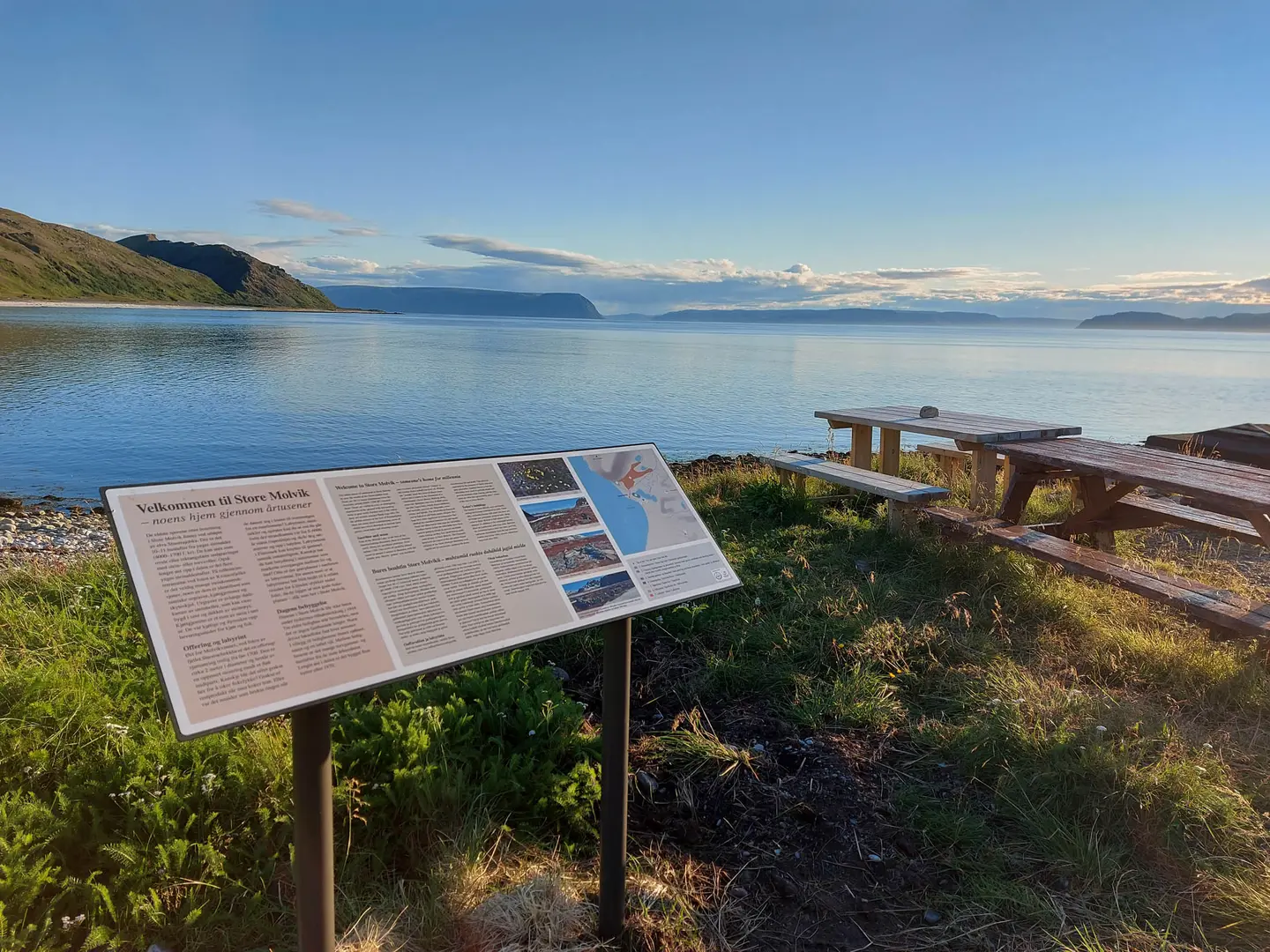 Et skilt med informasjon om området, med hav og fjell i bakgrunnen.