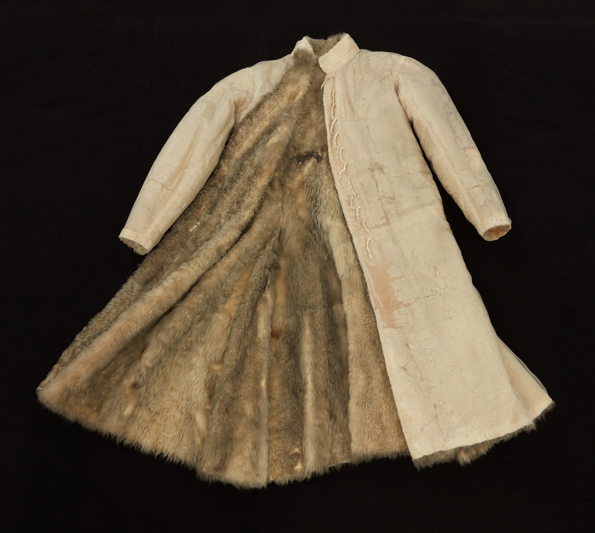 Skinnpäls tillhörande Gustav III:s daluniform. Långpäls av vita lammskinn, klädd med vit sidensars. Snitt som DM 110-111, men med smalt midjeband samt utan ärmuppslag. Knäppt åt höger med 12 små silverknappar och hyskor av skinn.