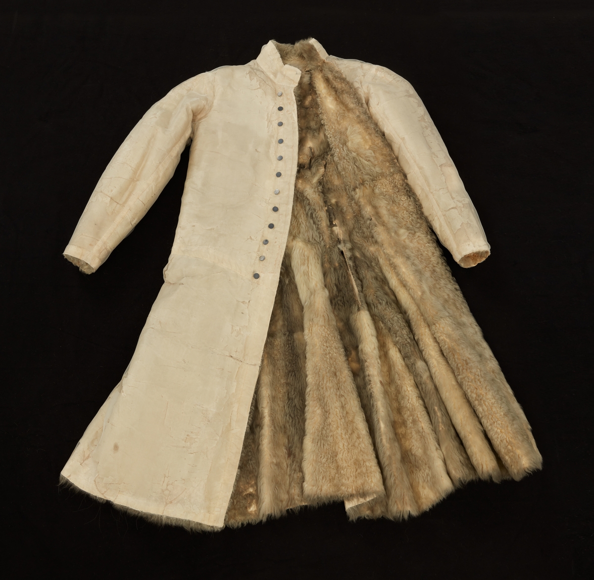 Skinnpäls tillhörande Gustav III:s daluniform. Långpäls av vita lammskinn, klädd med vit sidensars. Snitt som DM 110-111, men med smalt midjeband samt utan ärmuppslag. Knäppt åt höger med 12 små silverknappar och hyskor av skinn.