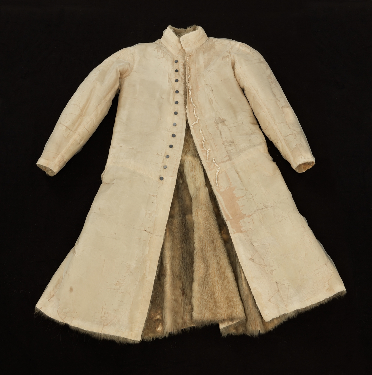Skinnpäls tillhörande Gustav III:s daluniform. Långpäls av vita lammskinn, klädd med vit sidensars. Snitt som DM 110-111, men med smalt midjeband samt utan ärmuppslag. Knäppt åt höger med 12 små silverknappar och hyskor av skinn.