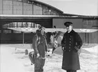 Två flygmilitärer står framför flygplan Ö 1 Tummeliten vid hangar på F 3 Malmen, vintertid, vid mitten av 1930-talet. En av dem är iklädd flygdräkt.