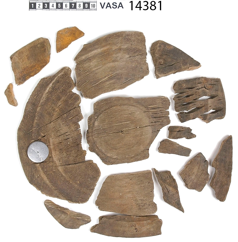 En träskål i delar.

Av delarna att döma har skålen varit plant samt försett med en svarvad fotring. På insidan av skålen, strax nedanför mynningskanten, har det löpt en dekorativ, svarvad linje. Mindre bitar av skålen saknas.

Delarna är mycket spruckna och mycket sköra.