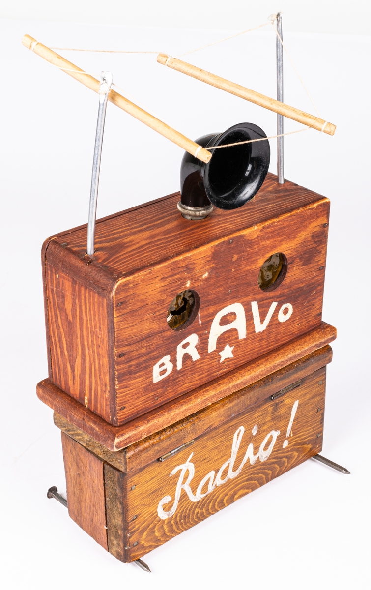 Radioapparat i flera delar. Trä. 
Radioapparat till dockteatern "Radiosabotören". Undre delen brunlaserad, skrivet "RADIO" med vit text. Övre delen har svart pastt ratt överst samt "antenner" i metall och snöre. Batteridriven.
Den andra delen är radion i "sprängt" utförande. Svartat och trasig. Plast tratten trasig bl.a