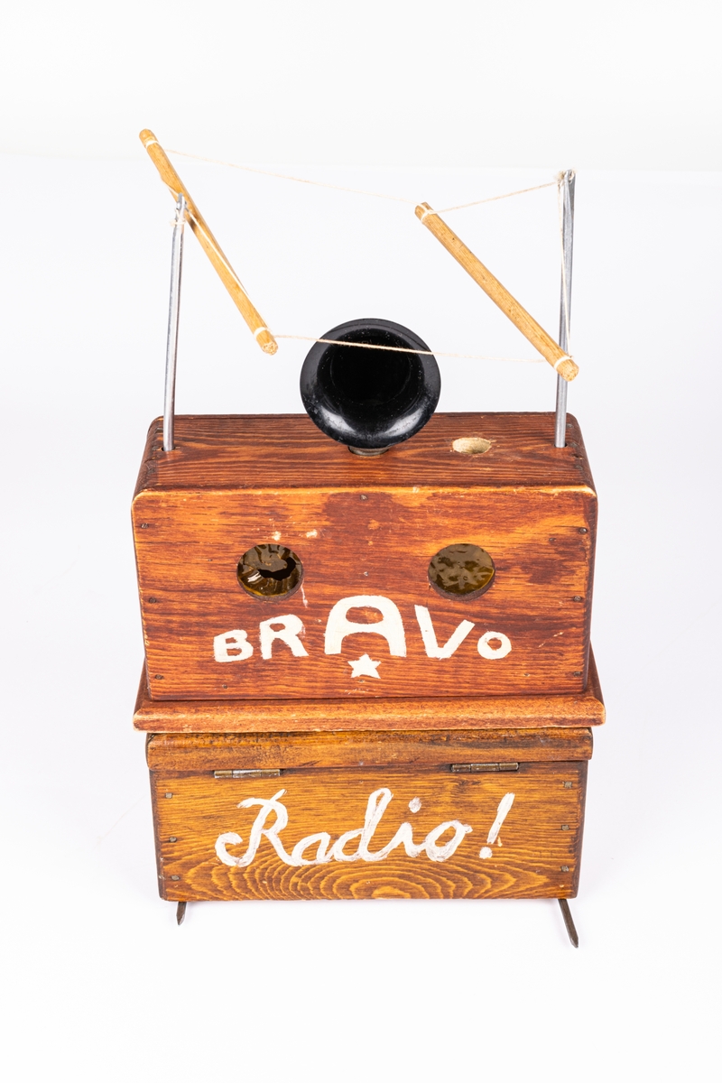 Radioapparat i flera delar. Trä. 
Radioapparat till dockteatern "Radiosabotören". Undre delen brunlaserad, skrivet "RADIO" med vit text. Övre delen har svart pastt ratt överst samt "antenner" i metall och snöre. Batteridriven.
Den andra delen är radion i "sprängt" utförande. Svartat och trasig. Plast tratten trasig bl.a