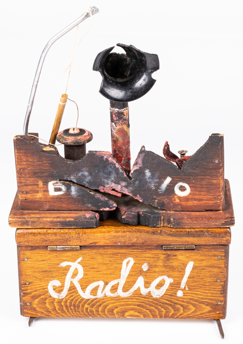 Radioapparat i flera delar. Trä. 
Radioapparat till dockteatern "Radiosabotören". Undre delen brunlaserad, skrivet "RADIO" med vit text. Övre delen har svart pastt ratt överst samt "antenner" i metall och snöre. Batteridriven.
Den andra delen är radion i "sprängt" utförande. Svartat och trasig. Plast tratten trasig bl.a