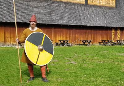 En mann i vikingdrakt, rød lue, spyd og et gult og grått rundt skjold står på en grønn plen - uten for en trebygning. Han ser mot kameraet.